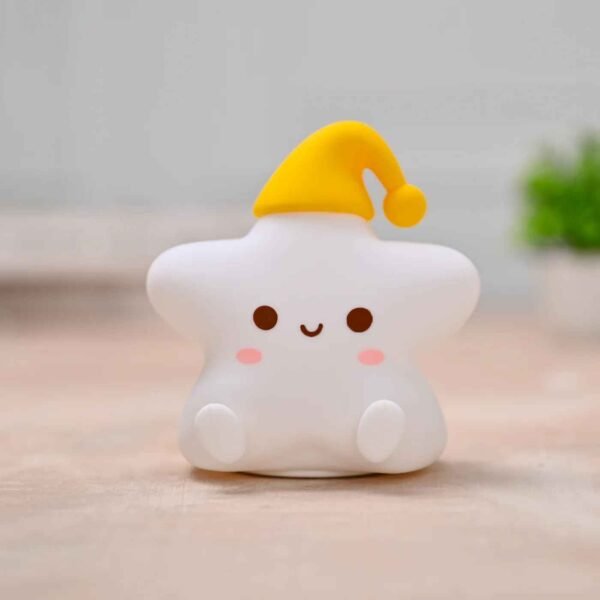 مصباح ليلي للأطفال على شكل نجمة بيضاء مبتسمة ترتدي قبعة نوم صفراء، موضوع على طاولة خشبية فاتحة.